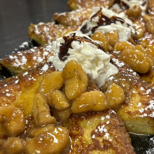 French toast topped with caramelized bananas, whipped cream, chocolate drizzle, and powdered sugar.
