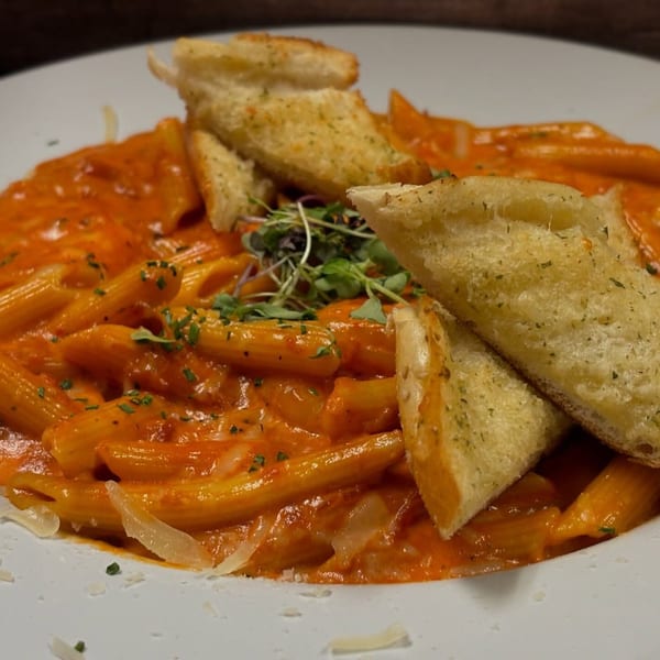 Penne pasta in a creamy red sauce, topped with microgreens and garlic bread on a white plate.