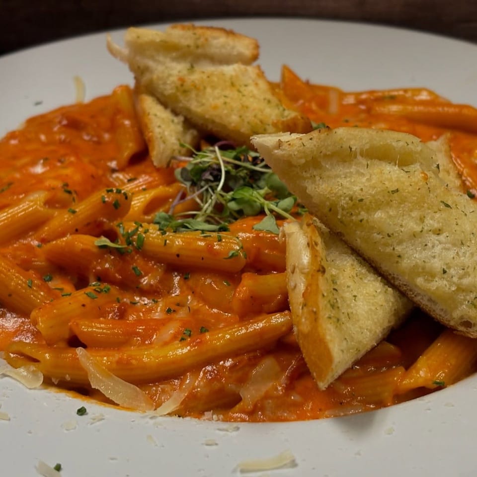 Penne pasta in a creamy red sauce, topped with microgreens and garlic bread on a white plate.